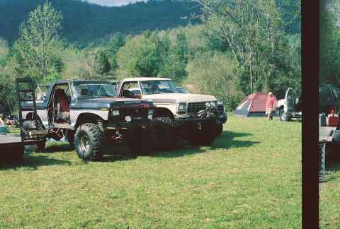 1978 Ford Bronco - Two modified 1978 Ford Broncos are parked on a grassy field with a tent and wooded hills in the background.