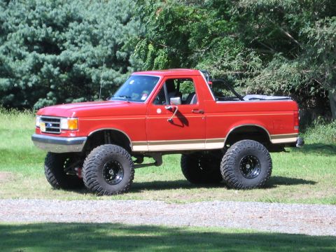 1990 Ford Bronco - Driver-side profile of a red Ford Bronco with tan lower two-tone stripe parked on a grass lawn beside a gravel drive, top and rear roof rem…
