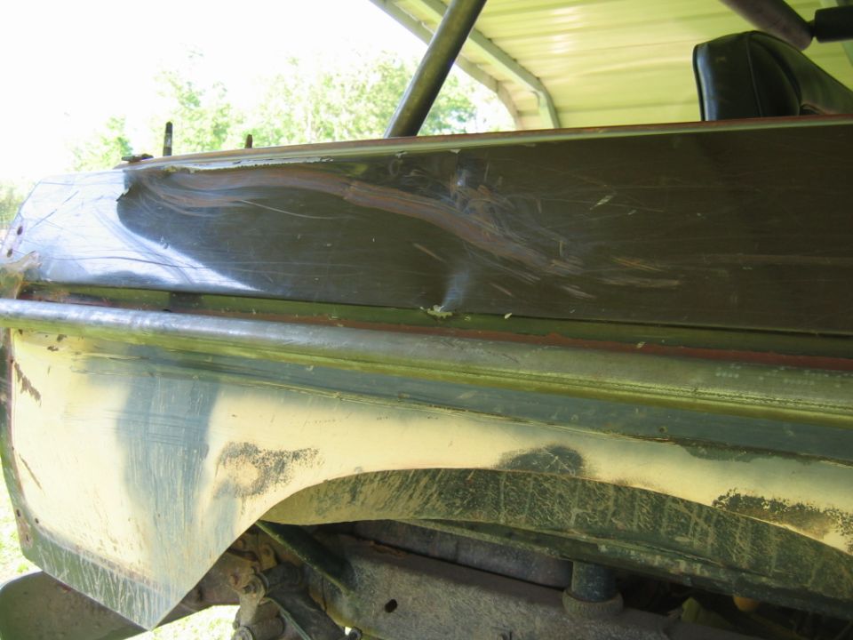 Damaged dark green vehicle body panel with scratches and a dent. Visible undercarriage and part of a seat