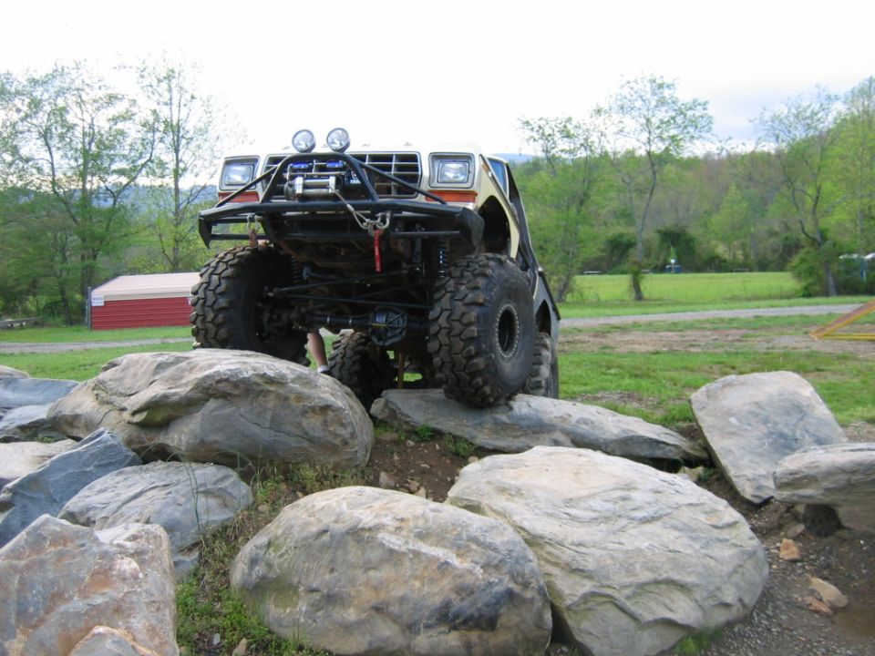 Modified off-road truck with large tires and a winch, climbing over rocks. Appears to be a Ford Bronco or F-series truck from the early 1980s