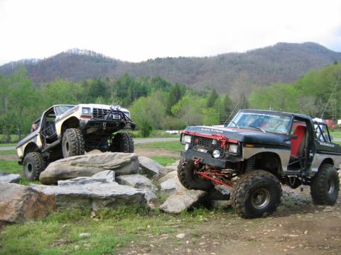 1992 Ford Mustang - Two heavily modified off-road trucks navigate a rocky terrain with a mountainous backdrop.