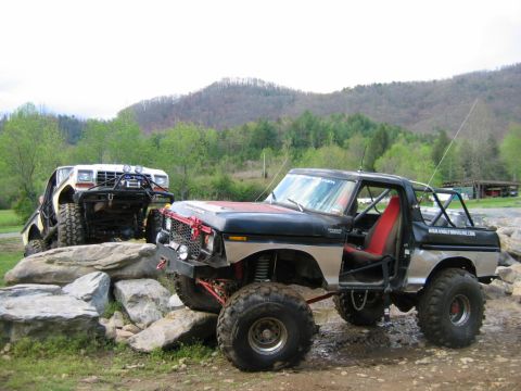1992 Ford Mustang - Two heavily modified off-road trucks are positioned on a rocky terrain with a wooded hillside in the background.