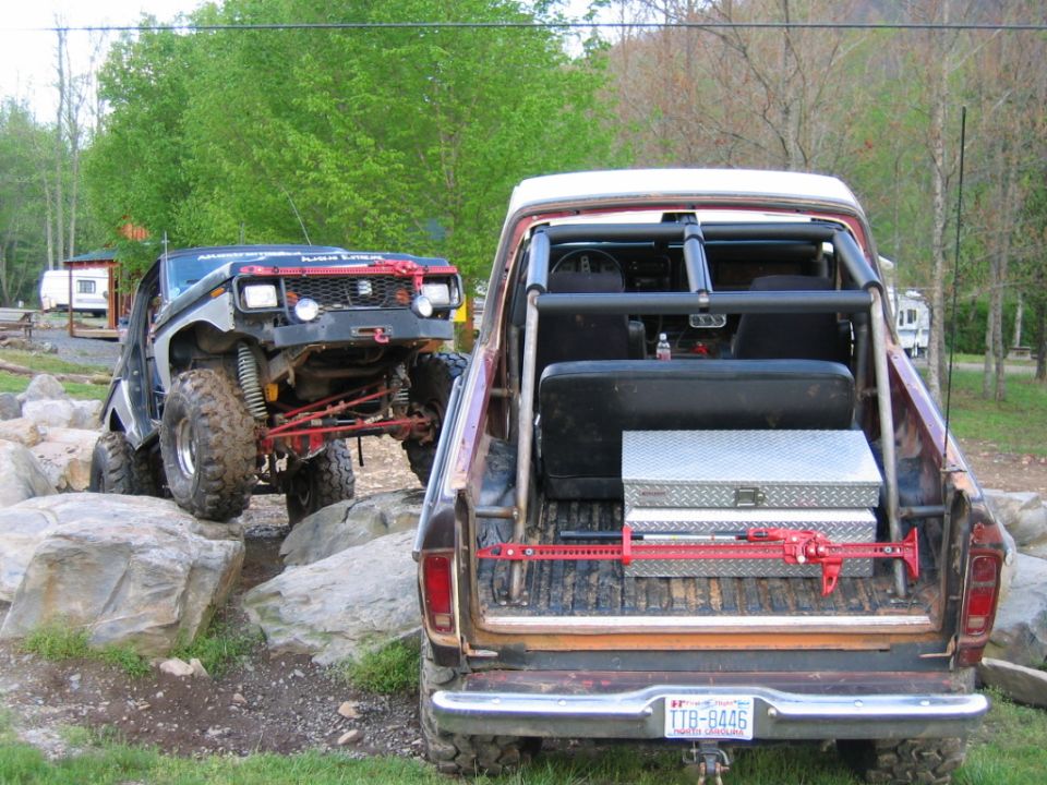 Two modified off-road trucks navigating a rocky terrain. One truck is in the foreground, viewed from the rear, showing its bed with a toolbox and jack. The other truck is on the left, climbing over rocks