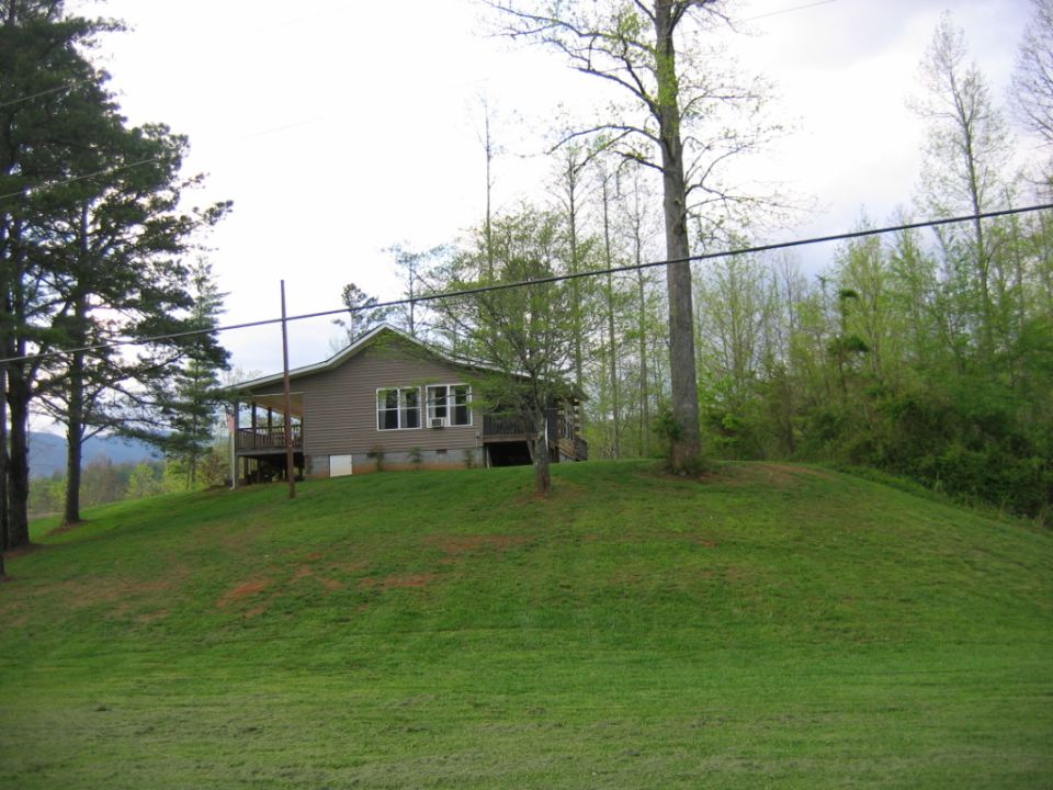 House with a porch sits on a green, grassy hill with trees in the background