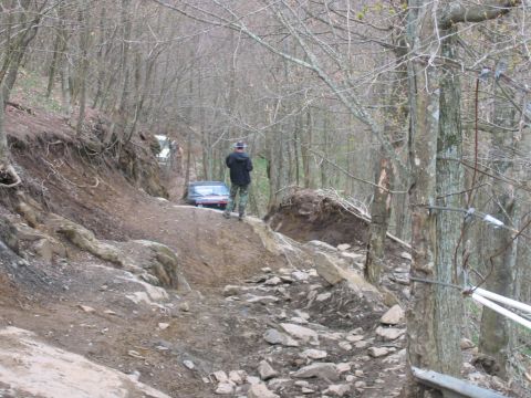 1992 Ford Mustang - A person stands watching as two vehicles traverse a challenging off-road path in a wooded environment.