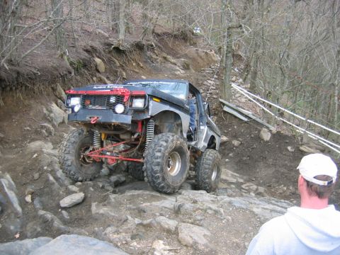 1992 Ford Mustang - A heavily modified off-road truck tackles a challenging rocky trail with a person watching.