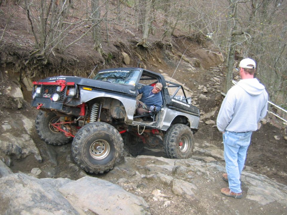 Modified pickup truck with large tires navigating a rocky trail, driver visible inside