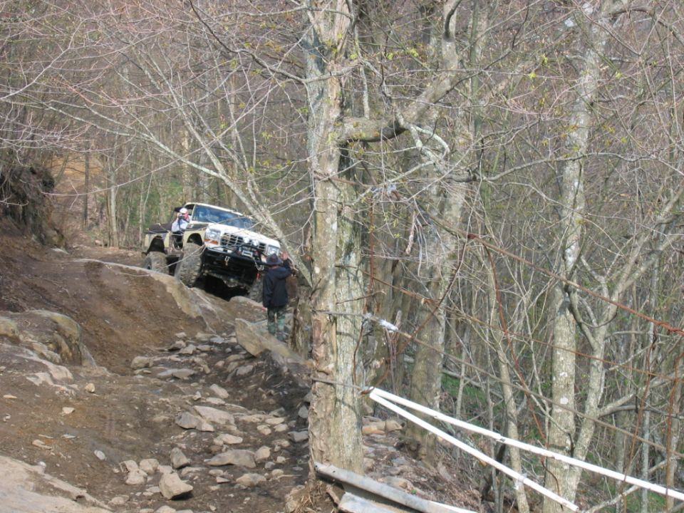 Off-road vehicle climbing a rocky trail with trees on either side. Two people are visible near the vehicle