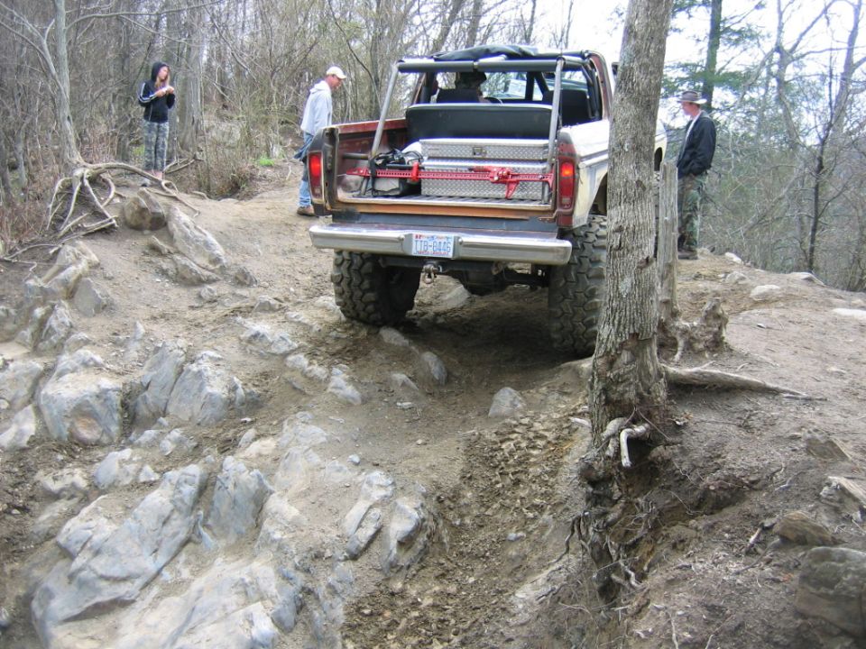 Off-road vehicle with large tires navigating a rocky trail. Toolbox and red straps visible in the open rear