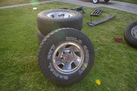 1986 Ford Bronco - Four Wild Country RVT radial tires and chrome wheels are stacked on a grassy lawn, with other vehicle parts visible in the background.