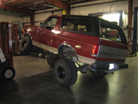 1990 Ford Bronco - Driver-side three-quarter rear view of a lifted red-and-tan full-size Ford Bronco with a black soft top, parked inside a metal-sided shop n…