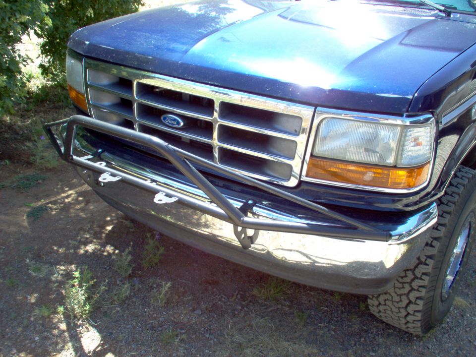 Blue Ford Bronco front end with chrome grille and aftermarket bumper. Tire and headlight visible