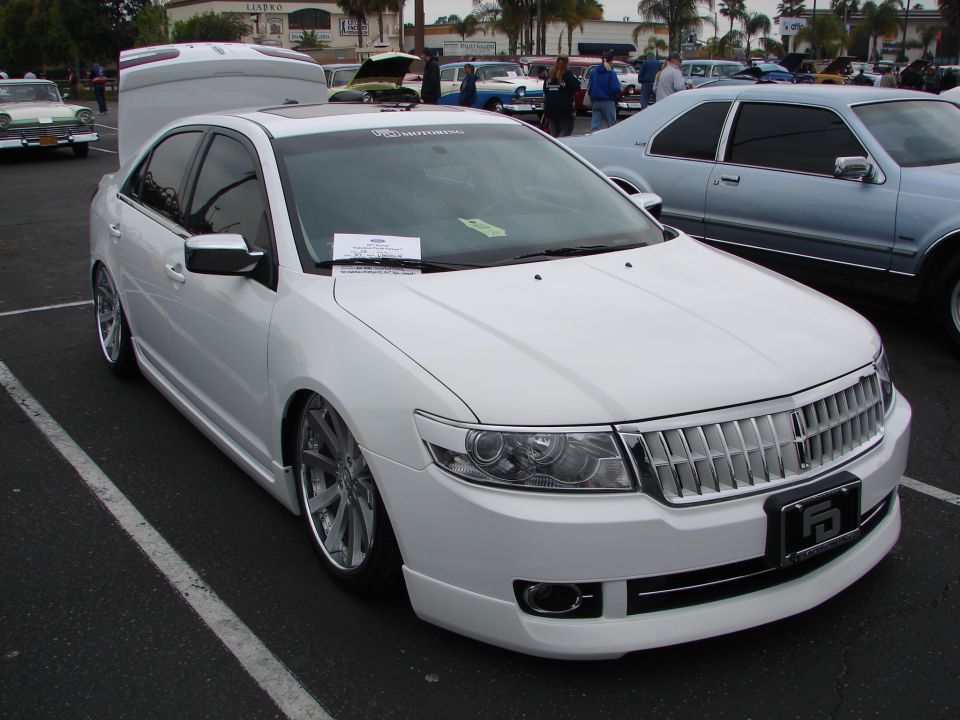 White sedan with chrome grille and custom wheels parked in a lot. Another car is visible to the right