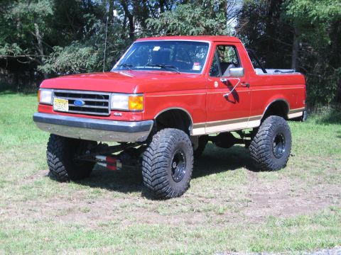 1990 Ford Bronco - Driver-side three-quarter front view of a lifted red full-size Ford Bronco with a tan lower stripe and the rear top removed, parked on gras…