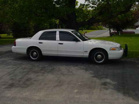 2002 Ford Crown Victoria - A white 2002 Ford Crown Victoria is parked outside on a paved surface with trees in the background.