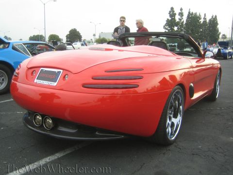1990 Ford Bronco - Rear view of a sleek red custom convertible concept car with dual exhaust and chrome wheels, two men standing behind it.