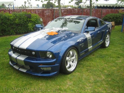 1990 Ford Bronco - blue Shelby Mustang coupe with silver racing stripes and chrome wheels parked on grass at an outdoor event.