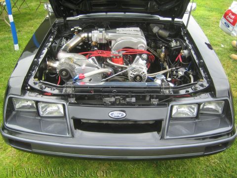 1990 Ford Bronco - Modified engine bay of a black Fox-body car with a Ford oval, polished intake and red plug wires, hood open on grass.