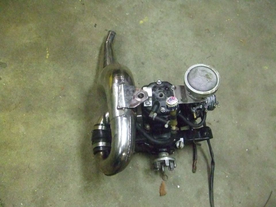 Two-stroke engine with a shiny exhaust pipe and air filter. It's sitting on a concrete floor
