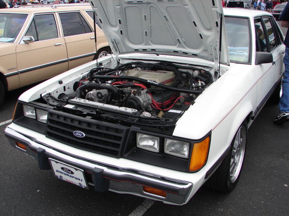 White Ford sedan with hood open showing the engine. Another beige sedan is parked next to it
