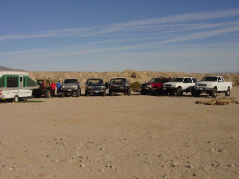 1994 Ford Bronco - A collection of trucks and a trailer are parked in a sandy, arid environment with hills in the background.