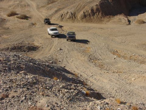 1994 Ford Bronco - Three trucks navigate a dusty, winding dirt road through a barren, mountainous desert landscape.