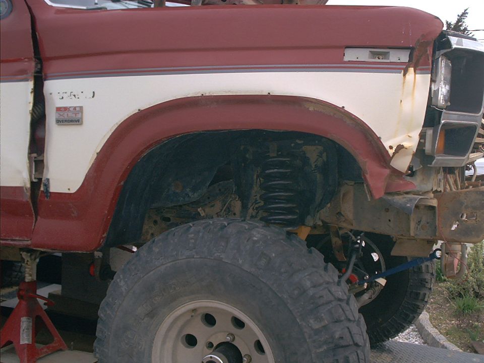 Red and white Ford Bronco fender, large tire, and suspension components. The front fender is damaged