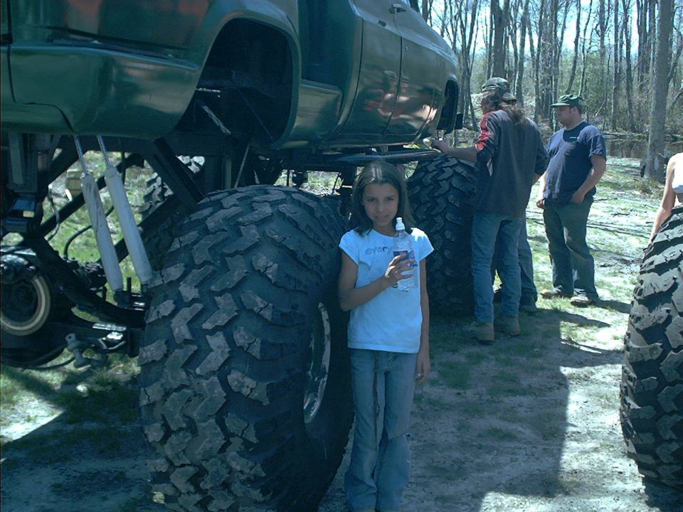 Lifted dark green pickup on huge mud tires with a child standing beside a rear tire for scale