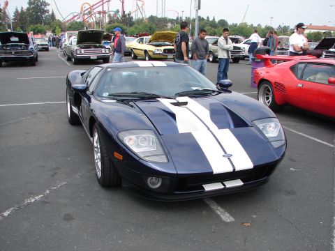 1992 Ford Taurus - A blue Ford GT with white racing stripes is parked in a lot, with other cars and people around.