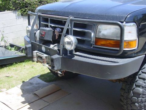 1994 Ford Bronco - Close-up of a dark blue Ford truck's aftermarket front winch bumper with an integrated black tube brush guard, diamond-plate top surface, a…