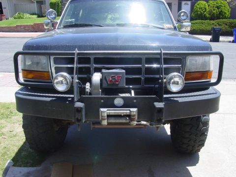1994 Ford Bronco - Head-on view of a dark green lifted Ford truck front end in a driveway, fitted with a heavy black winch bumper housing a Warn winch with fa…