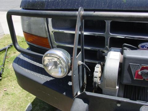 1994 Ford Bronco - Close-up of the front corner of a dark blue Ford truck fitted with a black tube grille guard and a diamond-plate winch bumper, with a chrom…