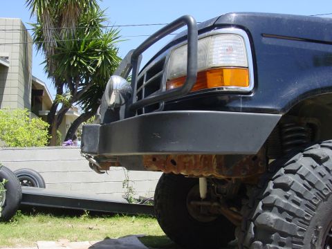1994 Ford Bronco - Low front-corner view of a lifted dark blue Ford truck in a yard, showing a black tube grille guard, chrome round driving light, heavy stee…