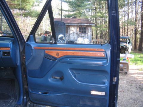 1979 Ford Bronco - Interior view of an open blue driver's door panel with wood-grain trim insert, manual door handle, armrest and lower carpeted section. Thro…