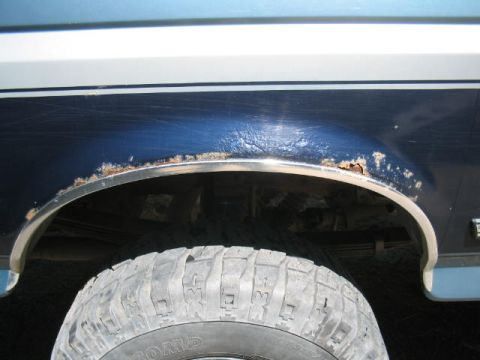 1979 Ford Bronco - Close-up of a dark blue rear wheel arch on a vehicle, showing significant rust and paint bubbling along the fender lip above a chrome wheel…