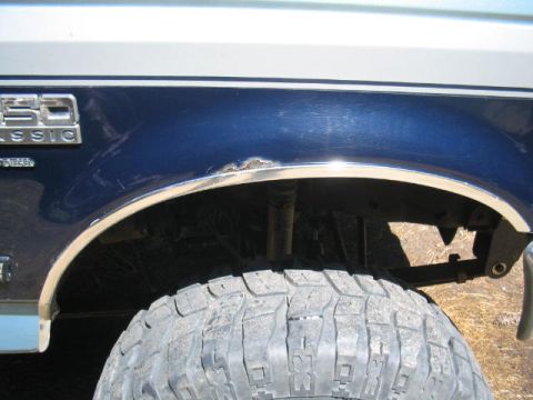 1979 Ford Bronco - Close-up of a dark blue Ford's rear fender area with chrome wheel-arch trim, showing a small area of rust damage at the top of the wheel op…