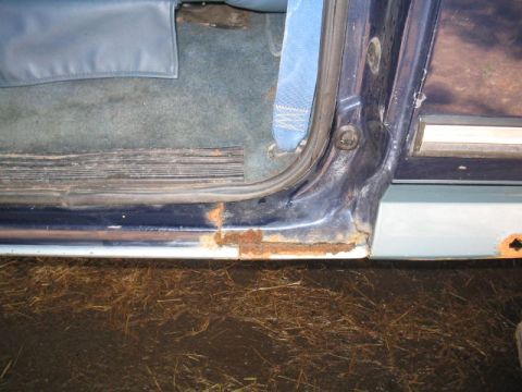 1979 Ford Bronco - Close-up of the lower door opening and rocker area of a dark blue vehicle, showing significant rust and corrosion along the sill and lower…