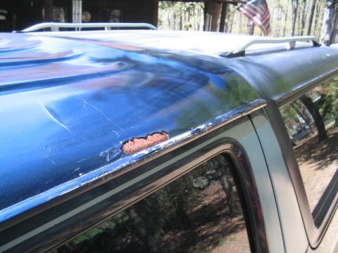 1979 Ford Bronco - Close-up of a blue SUV roof showing rust damage and paint bubbling along the drip rail edge above the side window, with a factory roof rack…