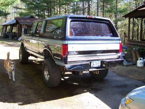 1979 Ford Bronco - Rear three-quarter view of a lifted two-tone blue and silver Ford SUV on large off-road tires, parked on a dirt driveway in a wooded settin…