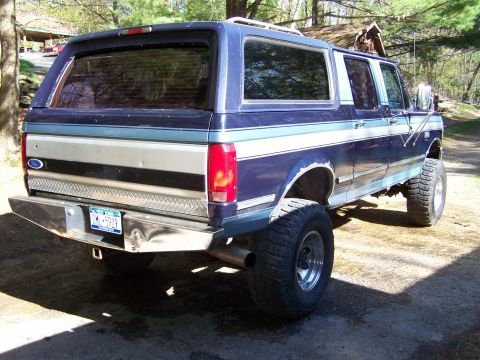 1979 Ford Bronco - Rear three-quarter view of a lifted two-tone blue and silver Ford truck with a matching camper shell/SUV conversion body, riding on large o…