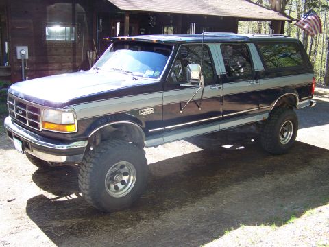 1979 Ford Bronco - A lifted two-tone black and silver Ford crew-cab conversion truck with a matching hardtop camper shell, photographed from the front-left th…