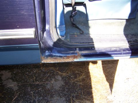 1979 Ford Bronco - Close-up of a dark blue vehicle's rocker panel and lower door sill area showing significant rust and paint bubbling along the seam. The ope…