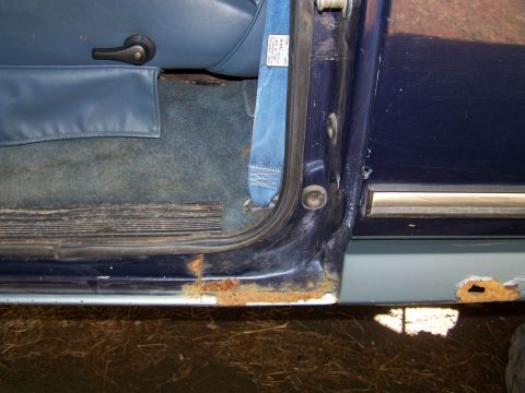 1979 Ford Bronco - Close-up of the driver's door sill and rocker area on a two-tone blue vehicle, showing significant rust and corrosion along the lower door…