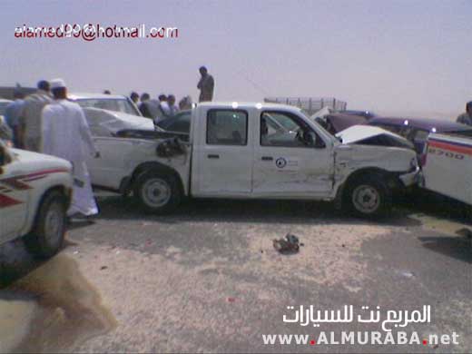 Damaged white pickup truck, front end crumpled, side dented, parked on a road with other vehicles