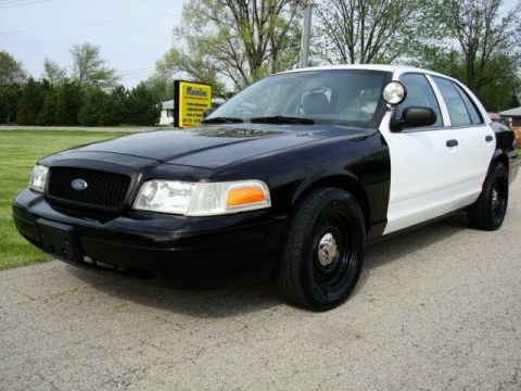 2003 Ford Crown Victoria - This two-tone police car is parked outside on a paved surface with grass and trees in the background.