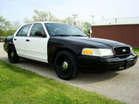 2003 Ford Crown Victoria - This 2003 Ford Crown Victoria police car is seen from the front passenger side, parked on a paved surface next to a grassy area.