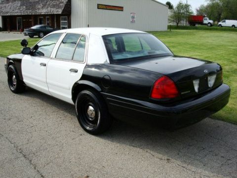 2003 Ford Crown Victoria - This two-tone police car is parked on a paved surface with a building and grassy area behind it.