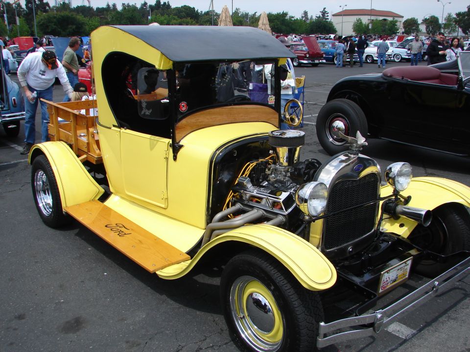 Yellow Ford hot rod truck with wooden bed and exposed engine. Another black hot rod is parked next to it