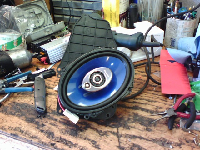 Car speaker with blue cone and silver tweeter, sitting on a workbench with tools like pliers and a utility knife