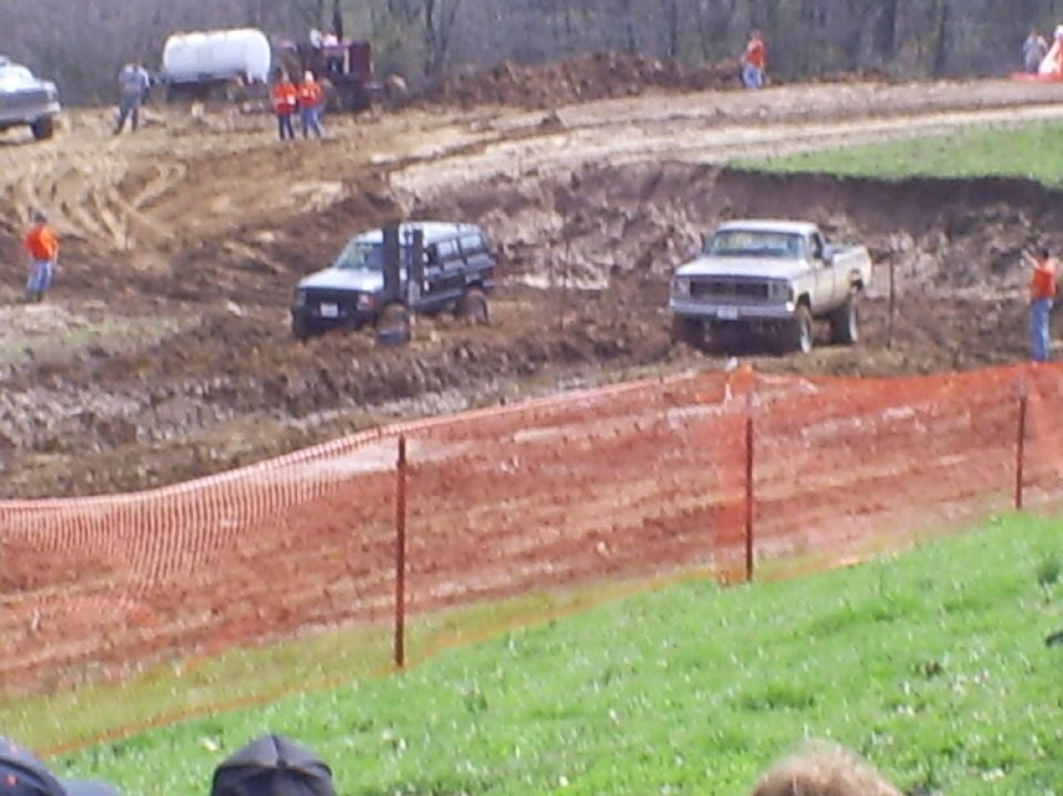 Two lifted 4x4 trucks in a muddy pit behind an orange safety fence at a mud bog event.
